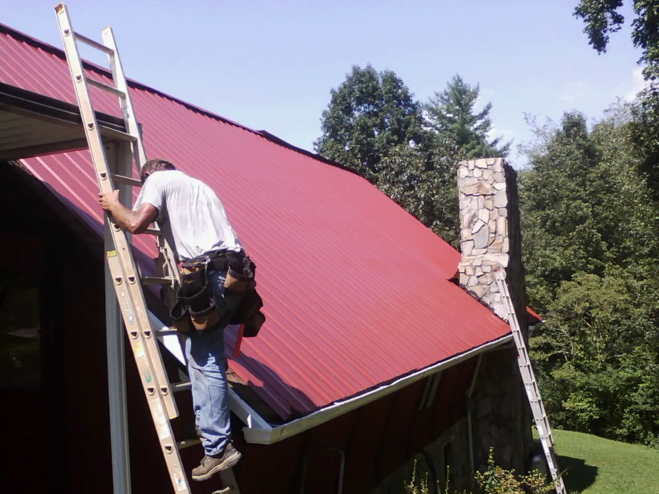 Roofer on a ladder beside a red metal roof and stone chimney — Soffit & Fascia Repair by Roofing Brothers in Lake Ozark